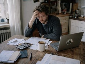 Freelancer aan keukentafel in de vroege ochtend omringd door papierwerk, facturen en een halflege koffiemok, met een natuurlijke uitdrukking van stille vermoeidheid